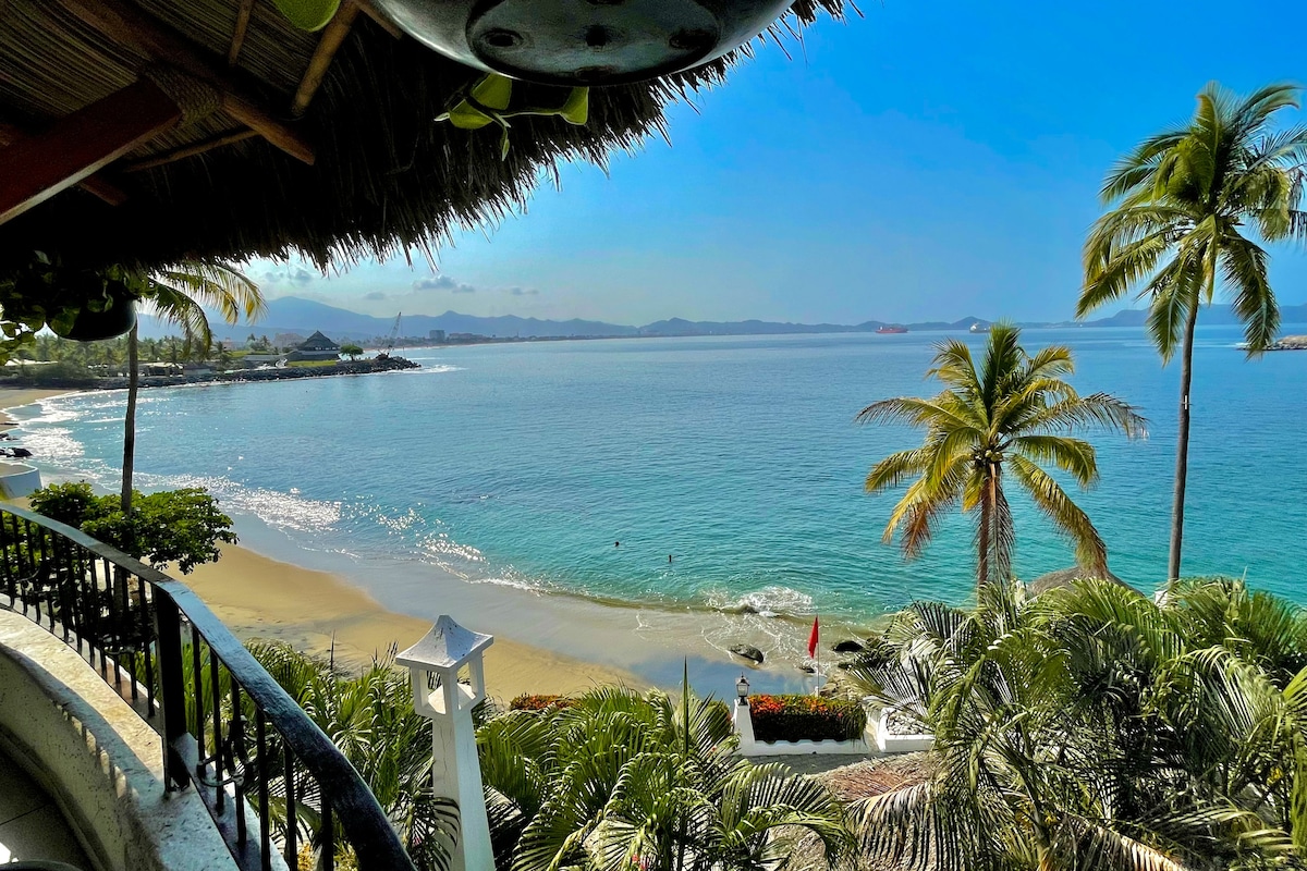 A wide view captures the serene ocean meeting the sandy beach, framed by palm trees. The calm turquoise waters reflect the sky, creating a tranquil scene. A lush landscape can be seen in the foreground, adding to the natural beauty of the coastal setting.
