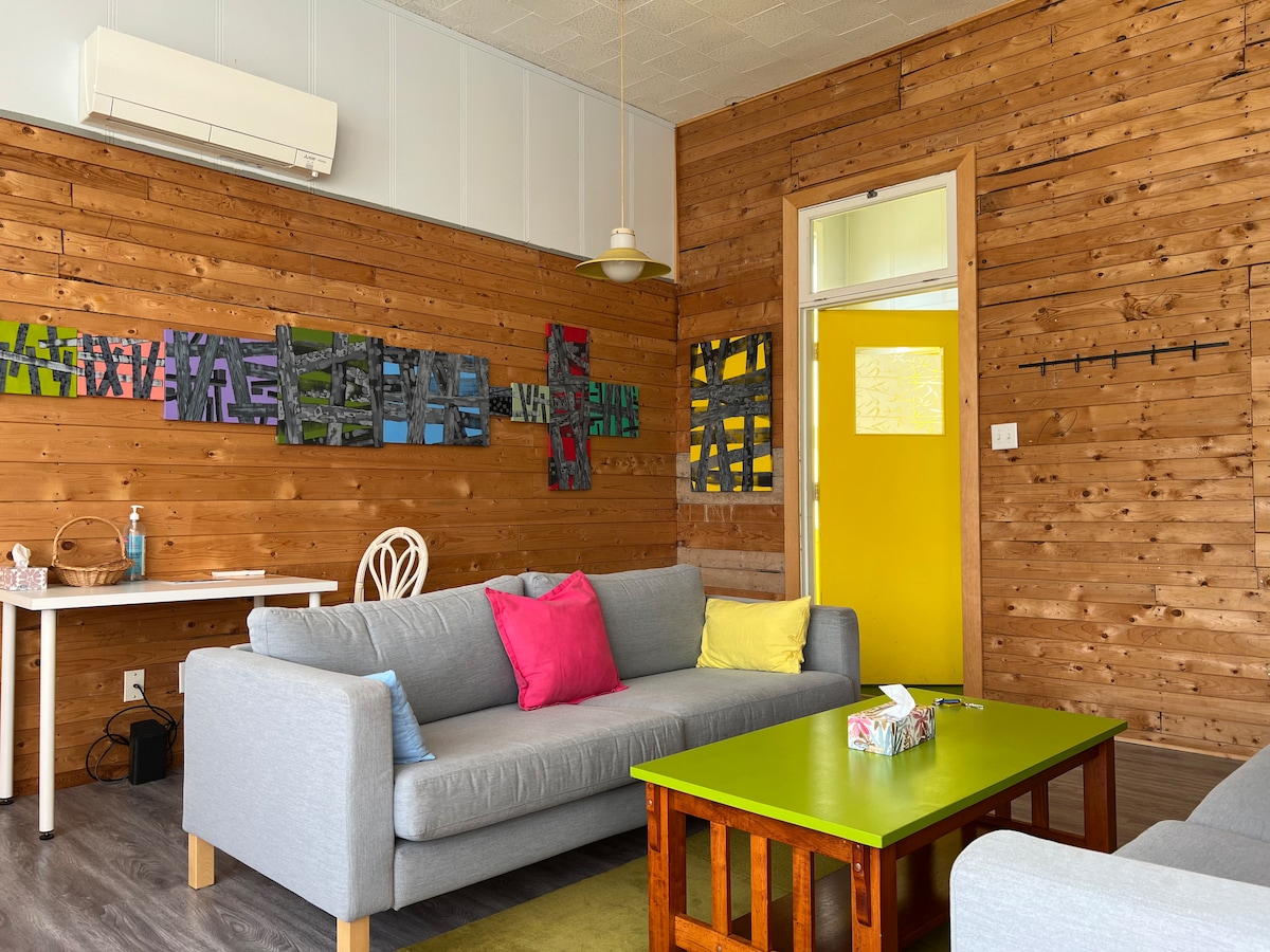 A comfortable living area features wooden walls that provide a warm backdrop. Two sofas, adorned with colorful cushions, are positioned around a vibrant coffee table. Wall art in various colors adds to the decor. A door painted in bright yellow leads to another space.