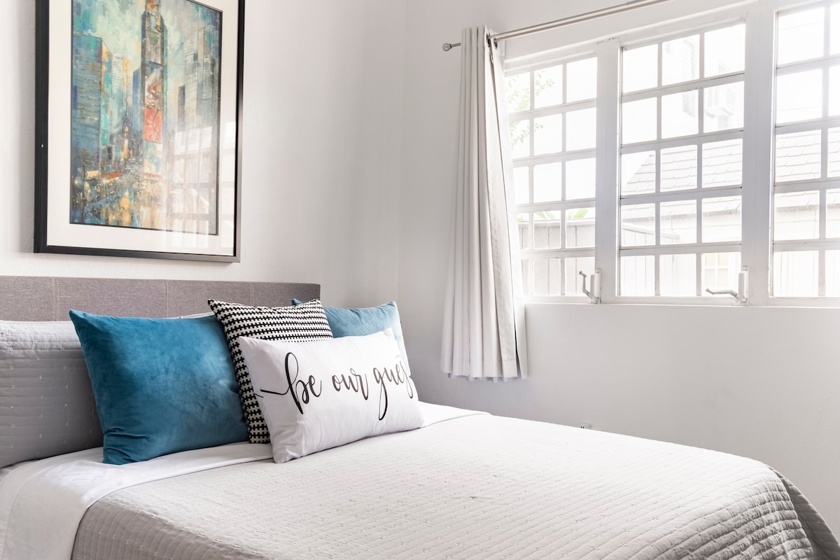 A well-lit bedroom features a spacious bed topped with an assortment of decorative pillows in various colors. Natural light filters through multiple windows with white curtains, enhancing the room's serene ambiance. A framed art piece is displayed above the bed.