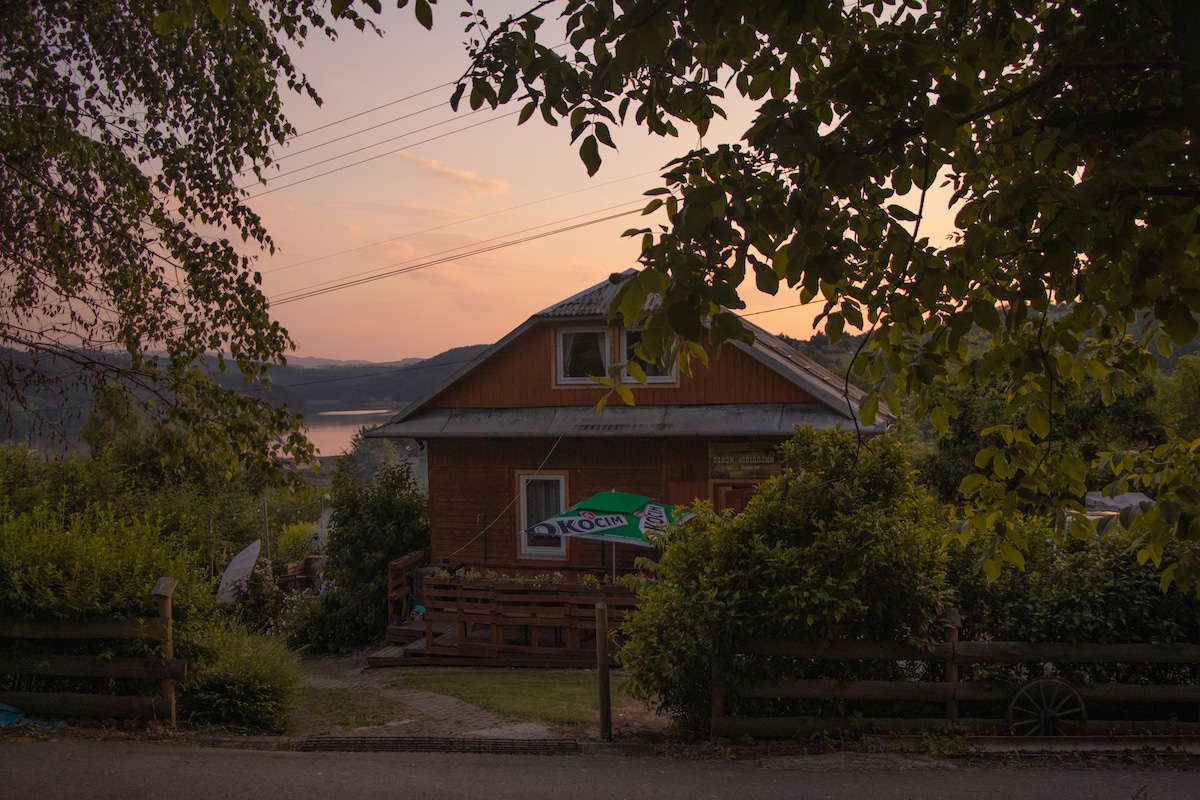 A charming wooden house is framed by lush greenery at sunset, with warm hues reflecting in the sky. The property features a deck area with outdoor seating and a sign for refreshments, creating an inviting appearance against the backdrop of nature.