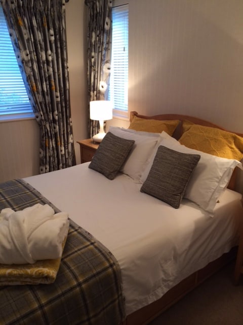 A double bedroom is present, featuring a neatly made bed with decorative pillows and a plaid throw. Natural light filters through window coverings, illuminating the soft hues of the space. A bedside lamp and wooden furniture add to the comfortable setting.