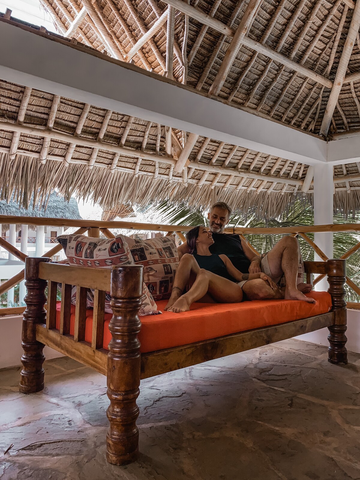 A spacious seating area is presented with a wooden frame and orange cushions, under a thatched roof. The floor is made of natural stone, and lush greenery is visible in the background, enhancing the relaxed ambiance of the space.