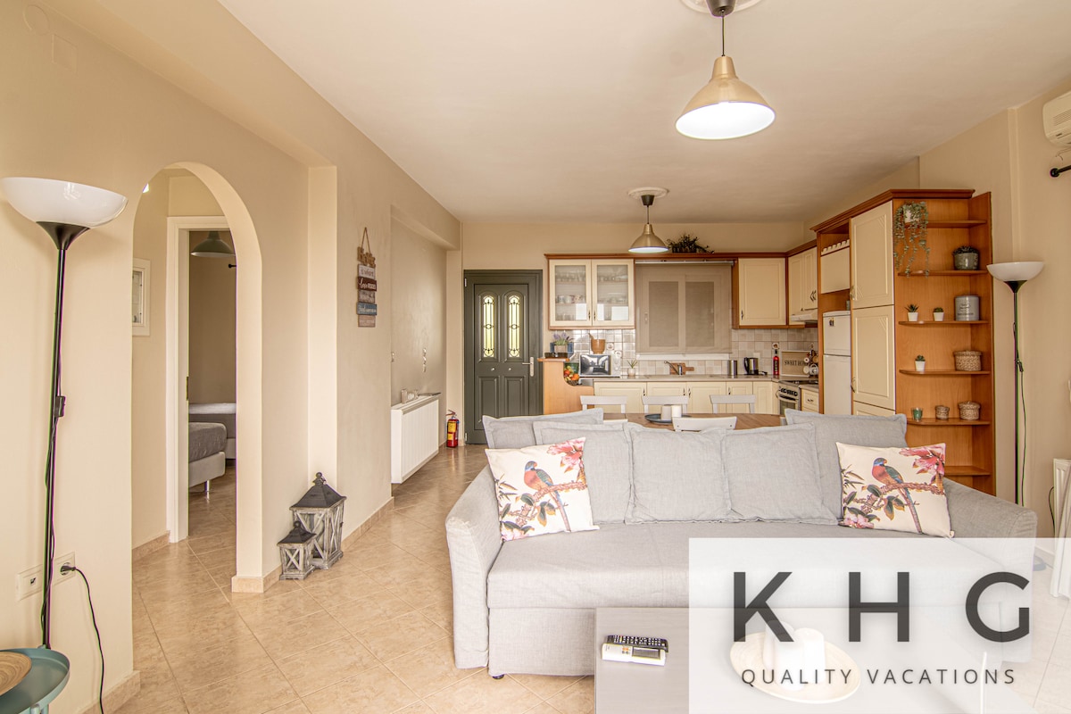 An open-plan living area features a light grey sofa adorned with decorative cushions, accompanied by a wooden coffee table. The space showcases modern kitchen amenities in the background, while an archway leads to a hallway. Natural light fills the room through multiple windows.