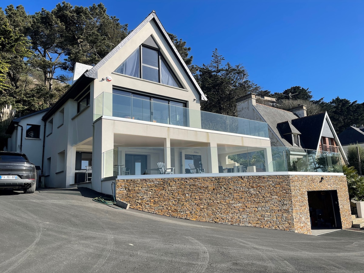 A modern villa is situated on a hillside, featuring a prominent glass-railed terrace that extends from the upper level. A stone facade complements the sleek design. The driveway leads to a covered parking area, and lush greenery frames the property.