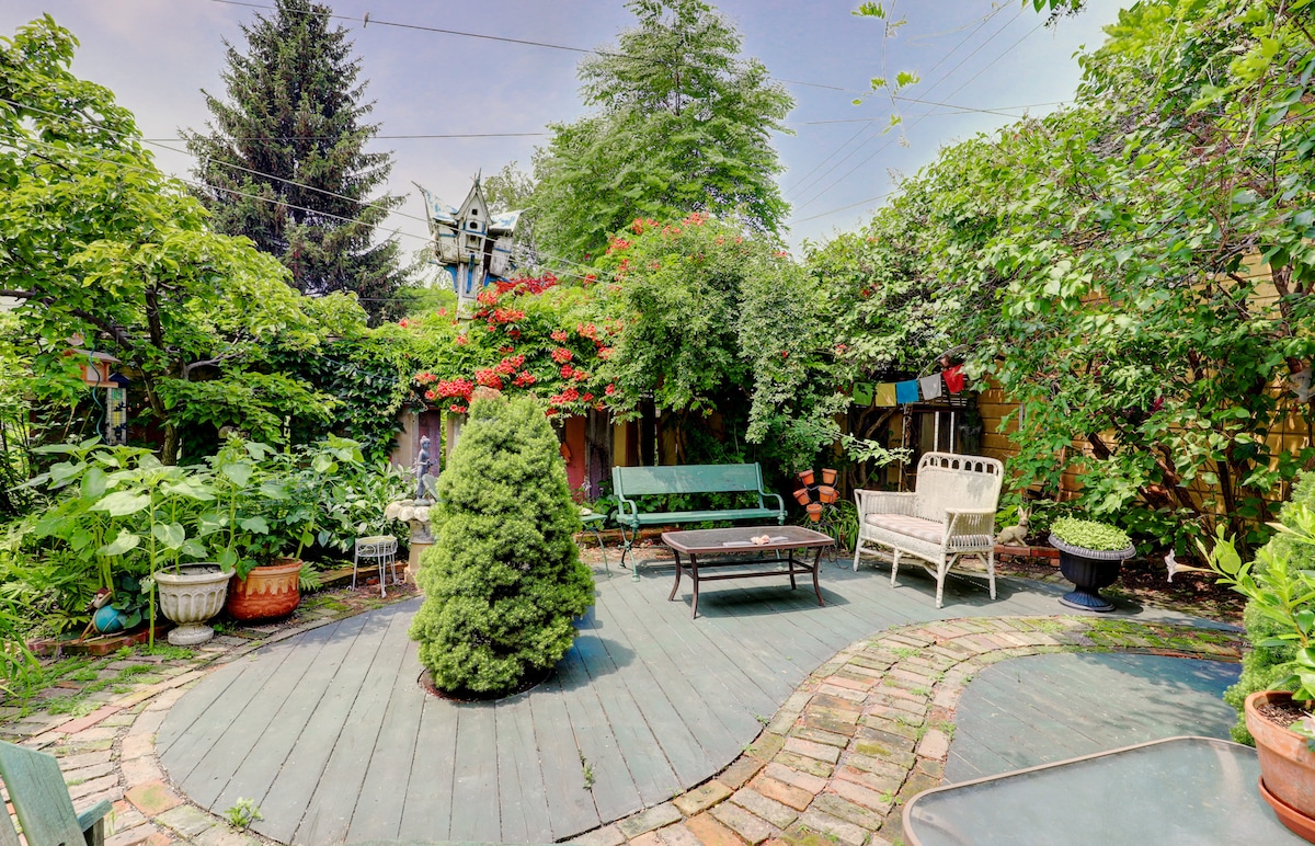 A serene garden area is framed by lush greenery and colorful flowers. Various seating options, including a wooden bench and a metal table, are surrounded by decorative planters. Stone pathways lead through the vibrant landscape, creating a welcoming outdoor space for relaxation.
