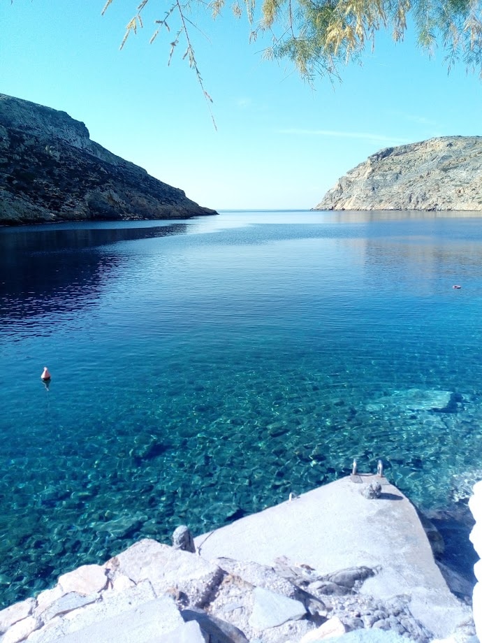 A calm sea glistens under clear blue skies, framed by rocky cliffs on either side. The water is crystal clear, revealing stones and marine life beneath. Floating buoys are visible in the distance, contributing to the serene coastal landscape.