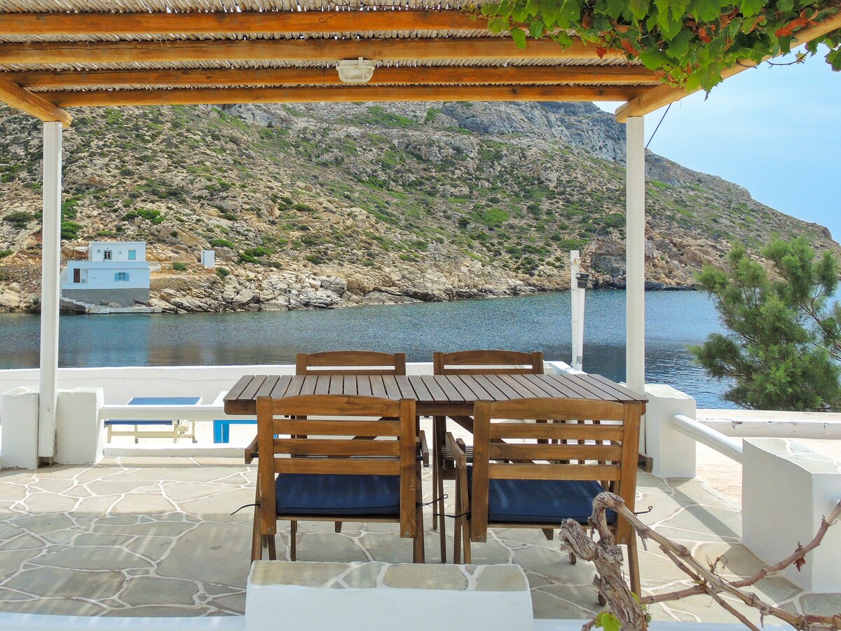 An outdoor terrace features a wooden dining set, including a table and chairs, overlooking the calm sea and rocky landscape. A canopy provides shade, while the natural stone flooring complements the serene coastal view.