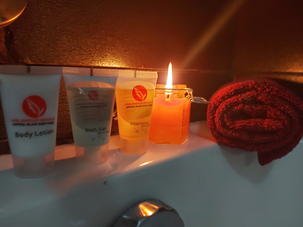 A selection of toiletries is arranged neatly beside a warm bath. Bottles of body lotion, bath gel, and shampoo are displayed alongside a lit candle, emitting a soft glow. A folded red towel is positioned nearby, adding a touch of comfort to the setting.