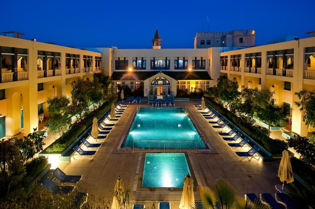 An aerial view captures the illuminated courtyard featuring a large swimming pool surrounded by lounge chairs. The warm lights accent the architecture of the building, highlighting the entrance and the trees that provide a serene atmosphere.