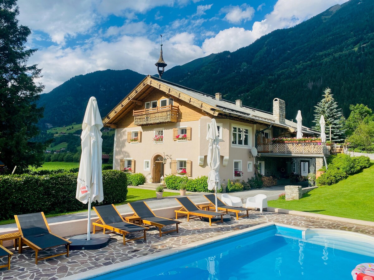 A charming building is showcased with upper-level balconies adorned with flower boxes. Sun loungers arranged neatly by a swimming pool invite relaxation, while the distant mountains provide a scenic backdrop. Umbrellas are positioned for sun protection, enhancing the outdoor leisure area.
