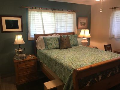A cozy bedroom is furnished with a spacious bed adorned in a green paisley-patterned quilt. Soft lighting is provided by two bedside lamps. Natural light filters through the sheer window coverings, while a wooden nightstand and decorative pieces enhance the room's inviting setup.