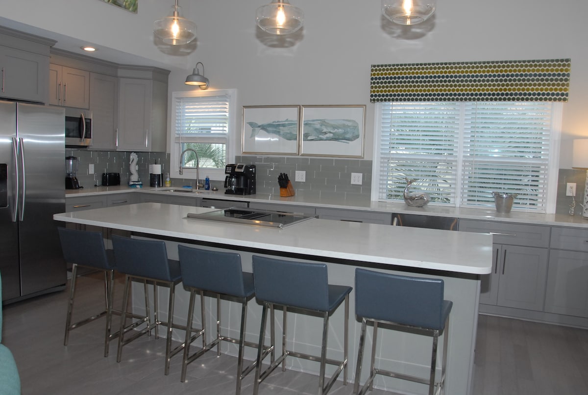 A modern kitchen is displayed with sleek grey cabinetry and a large island featuring bar seating for four. Stainless steel appliances, including a refrigerator and coffee maker, are visible. Bright natural light filters through the window dressed with patterned fabric, enhancing the airy feel of the space.
