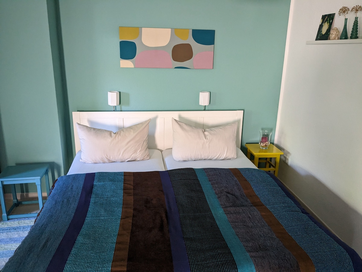 A cozy sleeping area is presented with a spacious bed adorned with a colorful striped blanket. Light-colored pillows rest neatly on the bed. An abstract wall art piece in pastel shades adds interest, while small side tables in contrasting colors complete the inviting look.