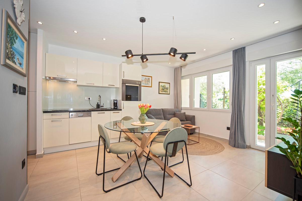 The open-plan living space features a modern kitchen with sleek cabinetry and appliances. A round dining table surrounded by four chairs is positioned near a comfortable sofa, with natural light streaming through large windows that overlook the garden.