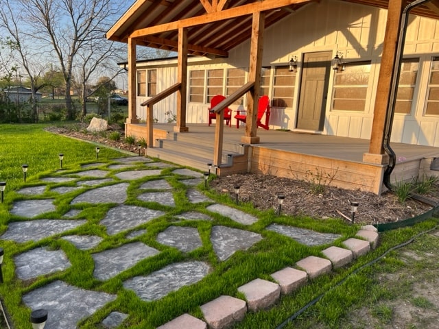 A well-maintained pathway of gray stone pavers is bordered by lush green grass, leading to the entrance of the farmhouse. Red chairs sit invitingly on the covered porch, framed by wooden beams and large front windows that allow for ample natural light.