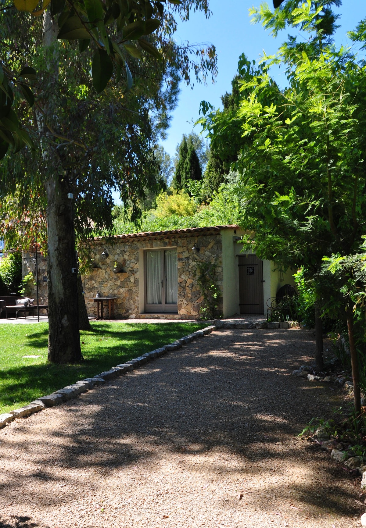 A charming cottage is surrounded by greenery, with a gravel path leading to its entrance. Tall trees and shrubs create a serene atmosphere. The cottage's stone facade complements the lush garden, inviting moments of relaxation in the natural setting.