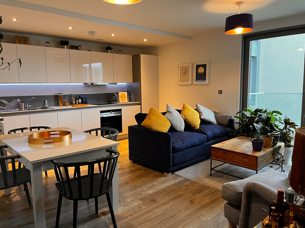 An open-plan kitchen and living space features modern cabinetry and a dining table set for four. A navy sofa with yellow pillows faces a glass balcony door, while greenery adds a touch of nature beside a wooden coffee table. Warm lighting creates a cozy ambiance.