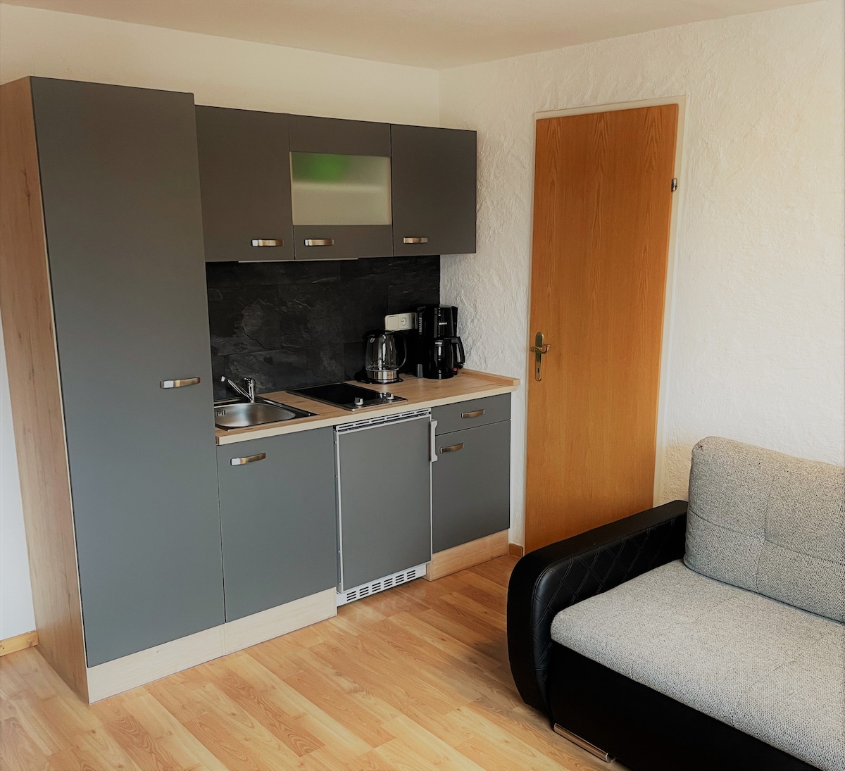 A compact kitchen area is equipped with modern gray cabinetry and a small kitchen countertop. Appliances include a refrigerator, kettle, coffee maker, and toaster. A seating area with a gray sofa is visible, providing a cozy spot for relaxation. A wooden door leads to another room.