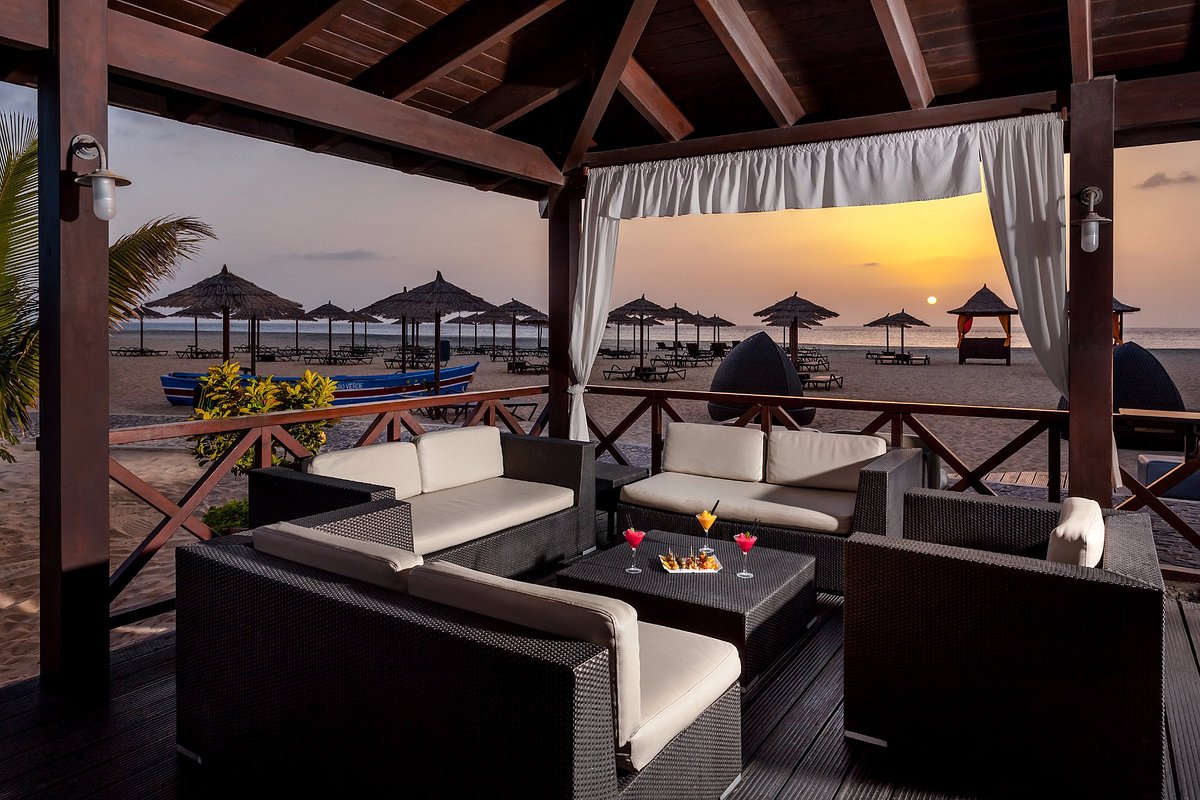 A shaded seating area features comfortable wicker furniture arranged around a central table. The view reveals a sunset over the beach, with silhouettes of thatched umbrellas in the background. Soft lighting complements the tranquil atmosphere, creating a welcoming space for relaxation.