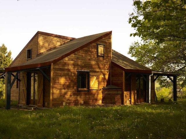 A wooden cabin is nestled amidst tall grass and trees, featuring a sloped roof and large windows. The porch front showcases an open area, providing a tranquil space to enjoy the natural surroundings.