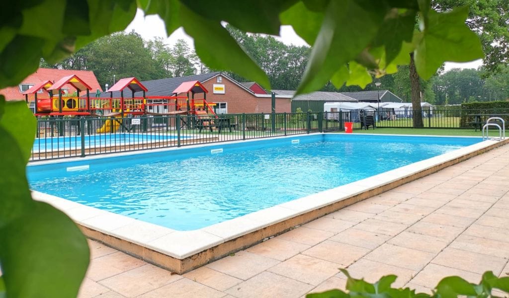 A fenced swimming pool is visible, featuring clear blue water and surrounded by a tiled patio. In the background, a brightly colored playground is seen, alongside additional outdoor structures, all set within a green landscape.