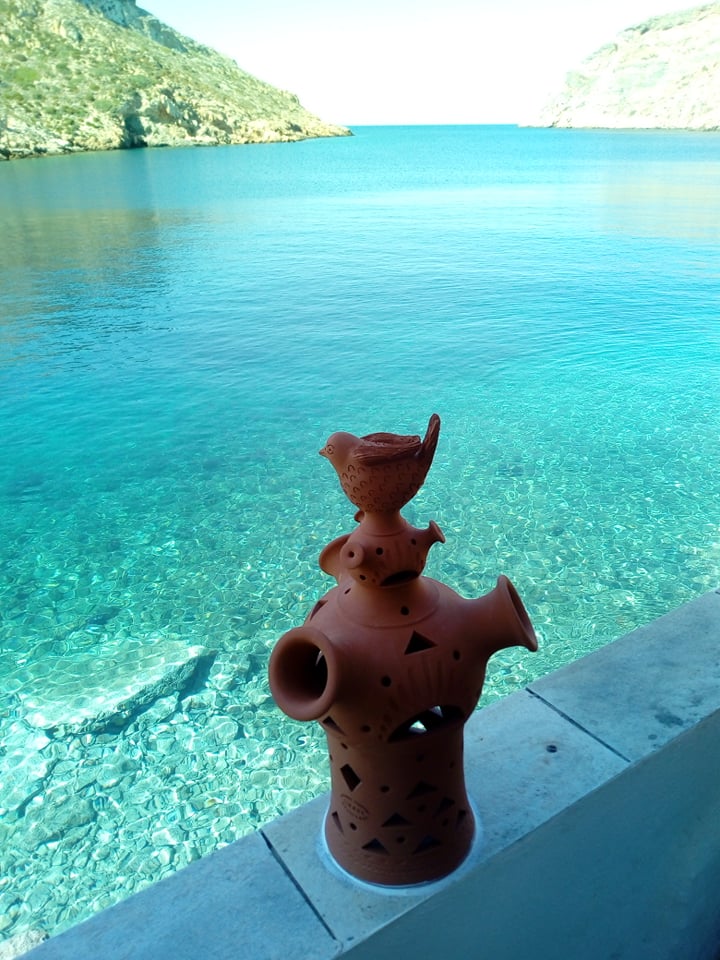 A terracotta pottery piece featuring intricate designs is positioned on a ledge. In the background, the clear blue water reflects light, bordered by cliffs that rise on either side, creating a serene coastal view.