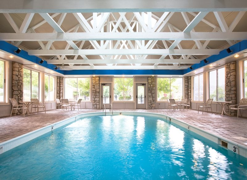 An indoor swimming pool is presented under a vaulted ceiling with exposed beams. Large windows allow natural light to fill the space, while lounge chairs are arranged around the pool area, creating an inviting atmosphere for relaxation.