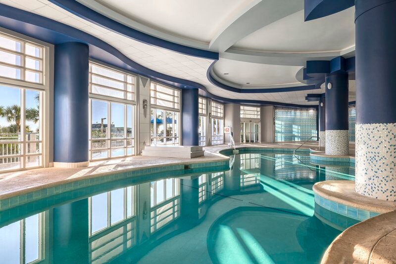 An indoor pool area is highlighted with smooth, curving walls painted in blue. Large windows allow natural light to flow in, reflecting off the calm water. Comfortable lounging areas with light-colored tiles are visible, enhancing the serene atmosphere.
