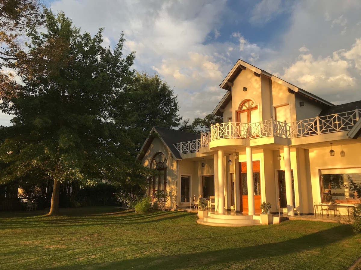 The exterior of a spacious house is showcased, featuring large windows and a welcoming entryway. A circular staircase leads to the entrance, surrounded by a well-kept lawn and mature trees. A balcony with decorative railing offers an inviting outdoor space.