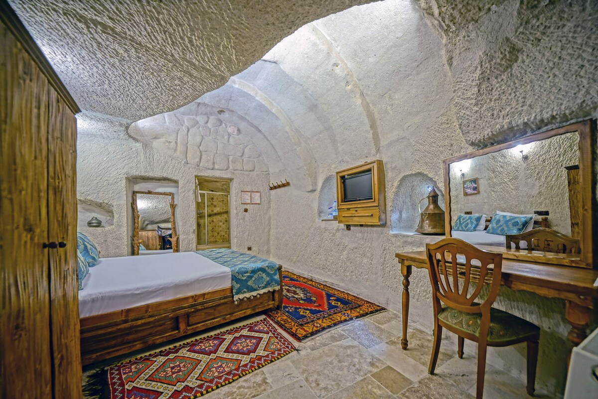 A charming cave room features naturally carved stone walls and an arched ceiling. A spacious bed is paired with a wooden wardrobe and a desk. A mirror is positioned above the desk, while a colorful rug adds warmth to the stone floor.
