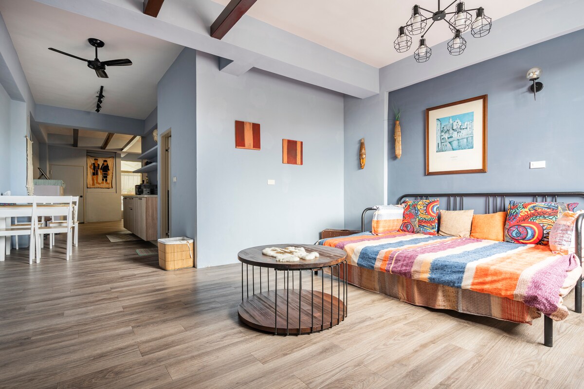 A spacious living area is furnished with a patterned sofa adorned with colorful pillows and a round coffee table. Light wood flooring complements the room, while artwork and soft lighting enhance the ambiance. A dining area and mini kitchen are visible in the background.