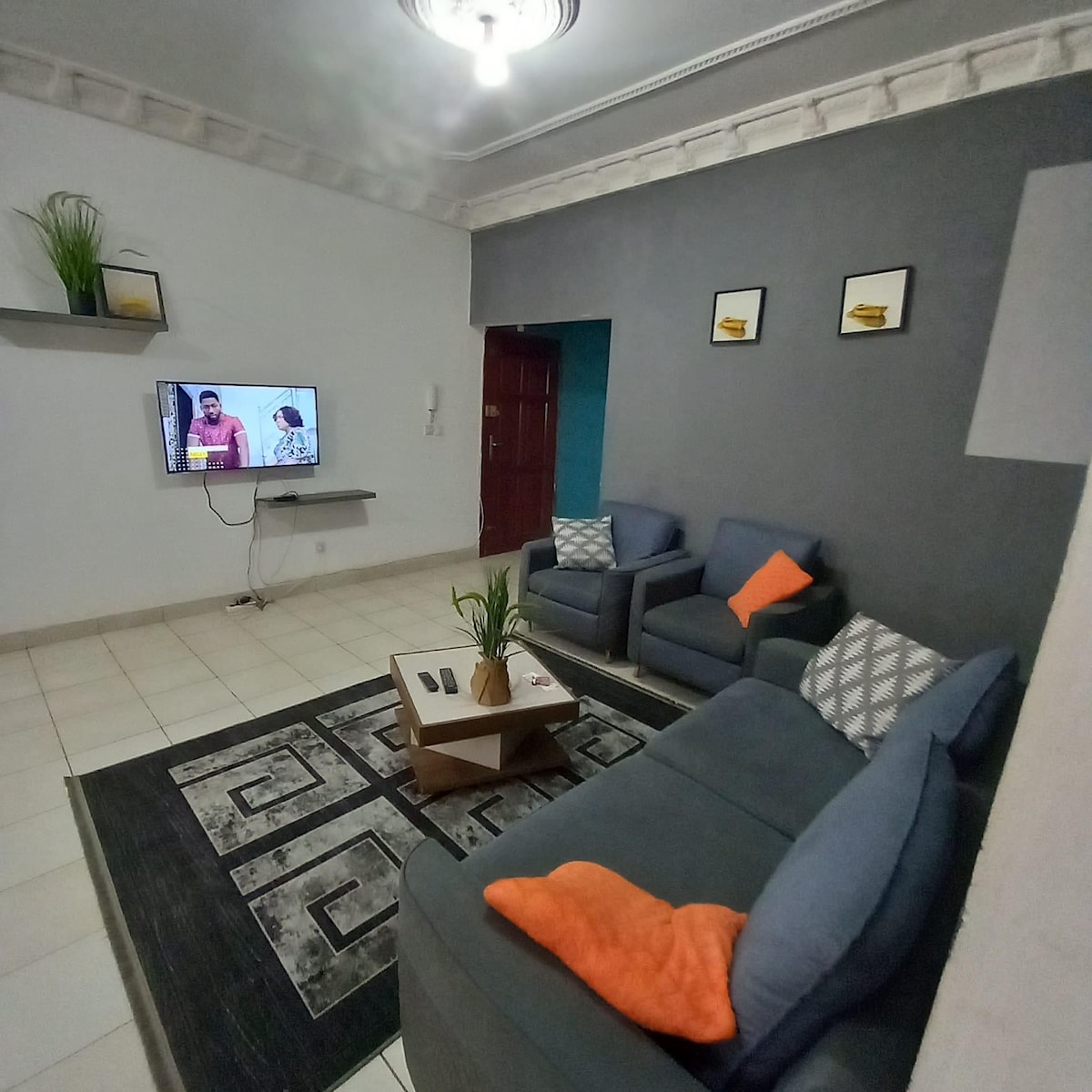 A modern living area features a comfortable sofa set with decorative cushions in vibrant colors. A flat-screen television is mounted on the wall opposite a light area rug that adds texture to the tiled floor. Greenery is present on a shelf, enhancing the space's tranquility.