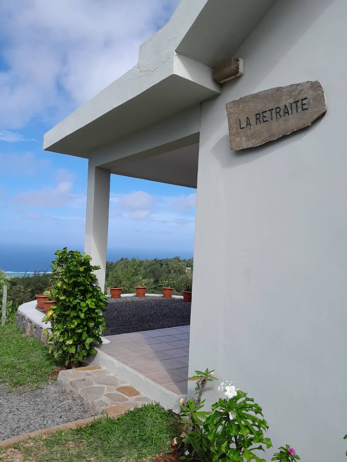 The exterior of the bungalow is highlighted, featuring a welcoming entrance with a stone sign reading 'La Retraite.' Potted plants are arranged along the pathway, and a clear sky serves as a backdrop, displaying a glimpse of the verdant surroundings and distant lagoon.