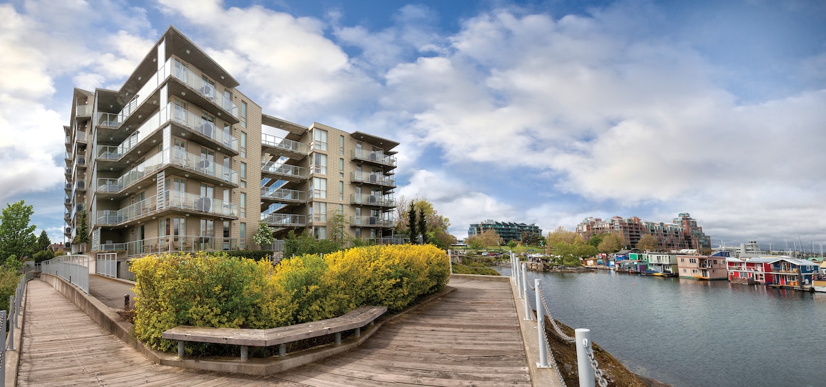 The exterior of the resort is shown, featuring a contemporary building with multiple balconies overlooking the water. Pathways lined with greenery lead towards the harbor, where colorful houseboats are visible in the distance, framed by a partly cloudy sky.