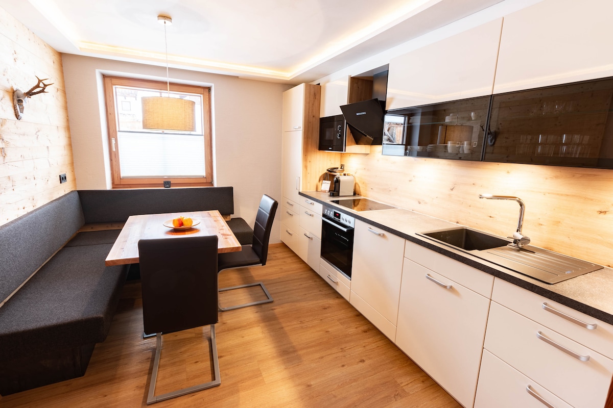 A modern kitchen area is presented with a sleek design featuring white cabinets and a black countertop. A wooden dining table is accompanied by black chairs, while a cozy bench seating is integrated along one side. Natural light filters through the window, enhancing the inviting atmosphere.