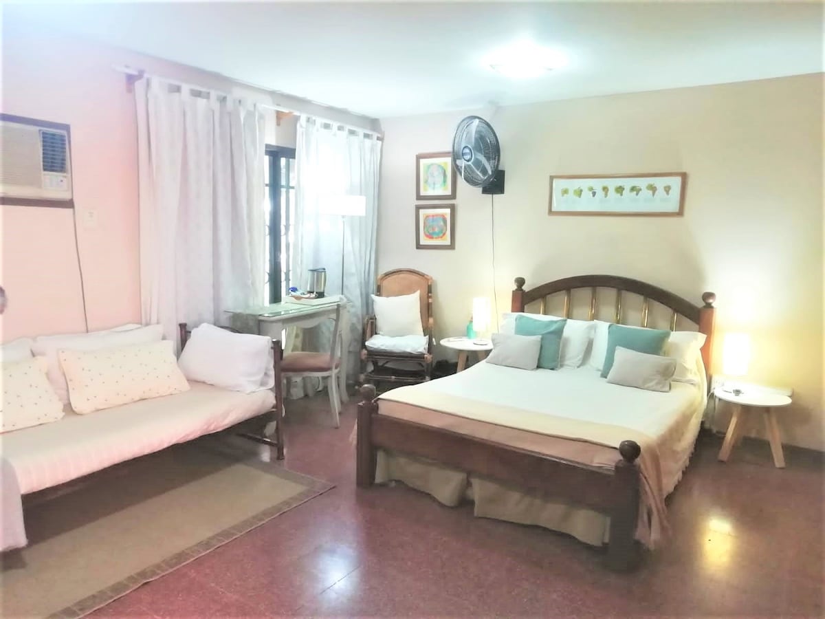 A spacious suite features a large bed with decorative pillows and a cozy seating area with a daybed. A small dining table is situated beside a window, complemented by a ceiling fan and wall-mounted air conditioning. Soft lighting creates a welcoming ambiance.
