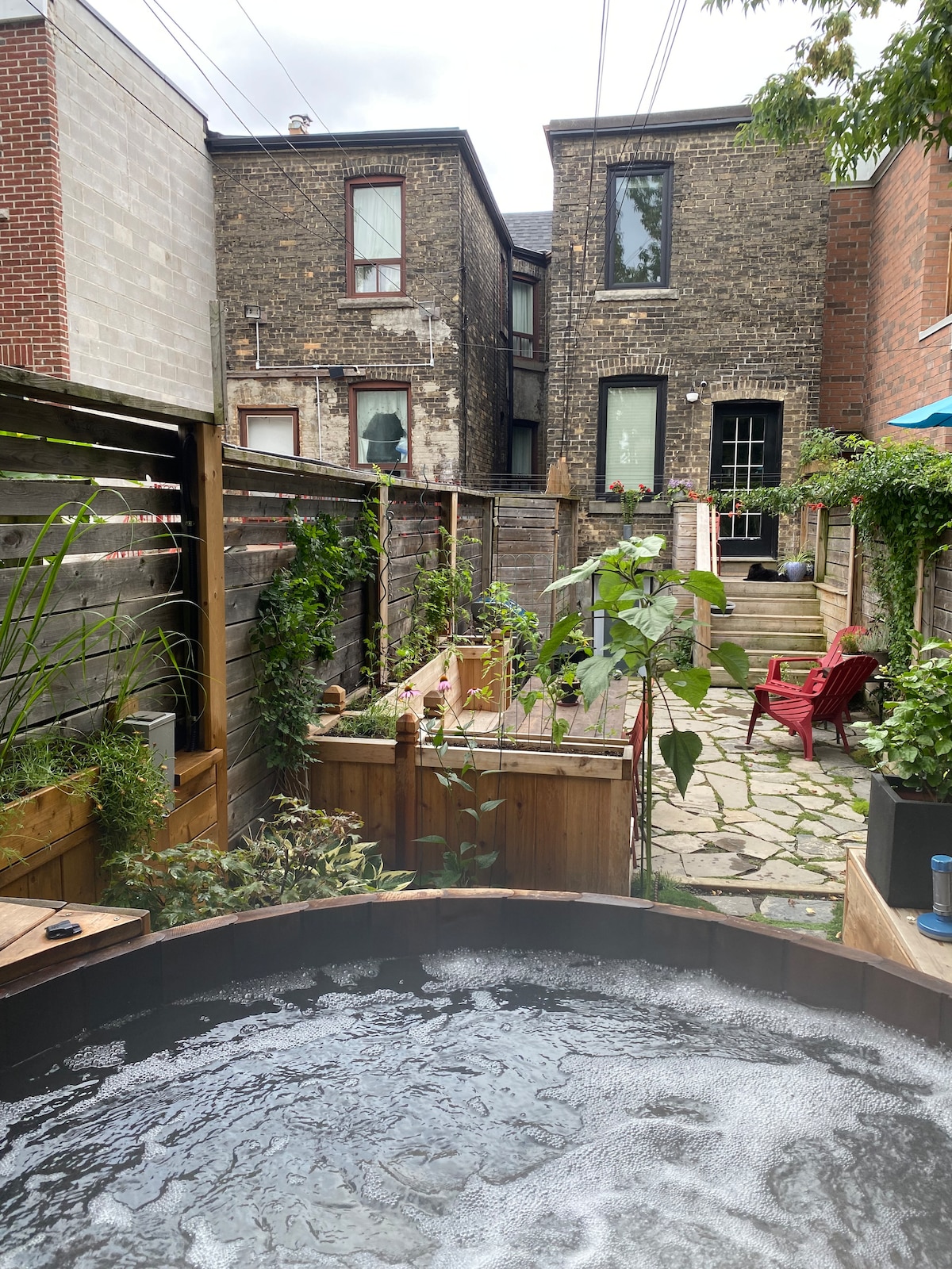 A relaxing backyard garden features a wooden hot tub with bubbling water, surrounded by lush greenery and wooden planters. A stone pathway leads to a set of stairs at the back, where a small patio area with a red chair is visible.