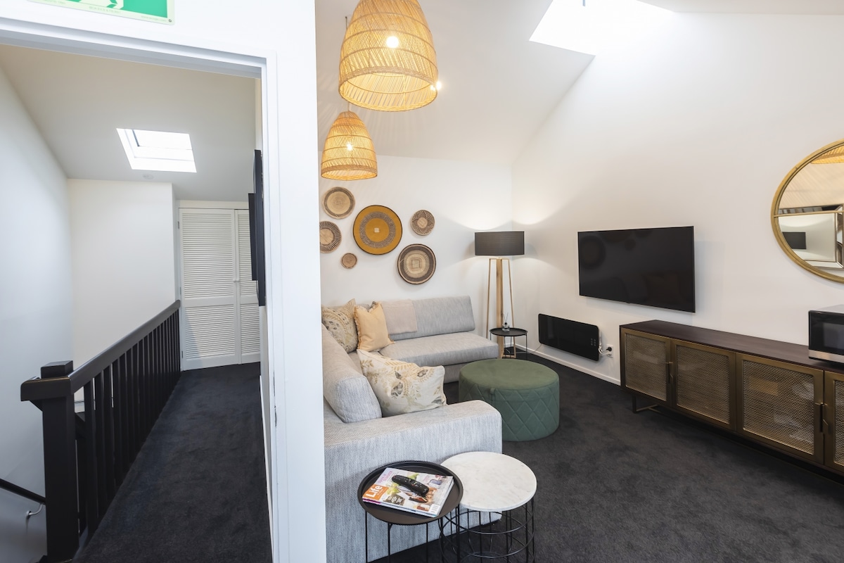 A bright living room is showcased, featuring a sectional sofa adorned with decorative pillows. A circular coffee table sits in front, alongside a small side table. A wall-mounted television is visible, with a sleek media console underneath. Natural light enters through a skylight.