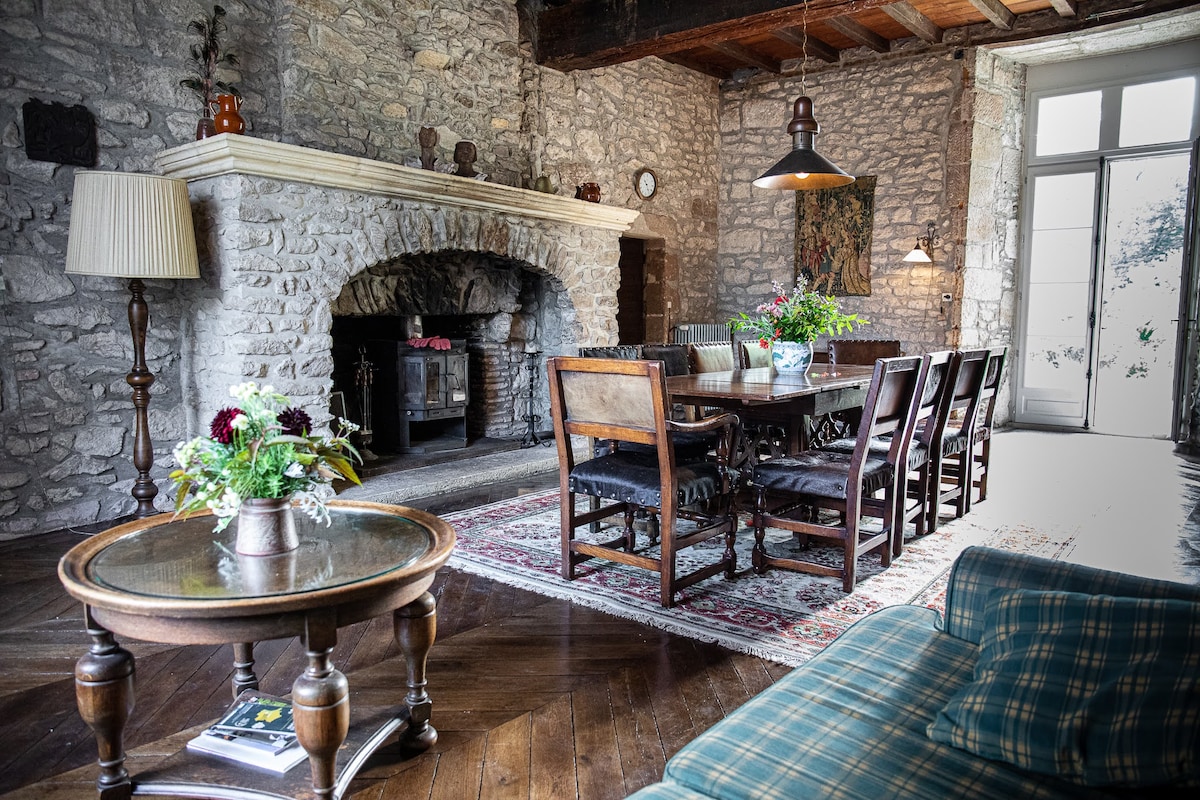 The living area features historical stone walls and a large open fireplace. A long wooden dining table is surrounded by eight chairs. A cozy seating area is across from the table, and natural light fills the space through large glass doors leading to the garden.