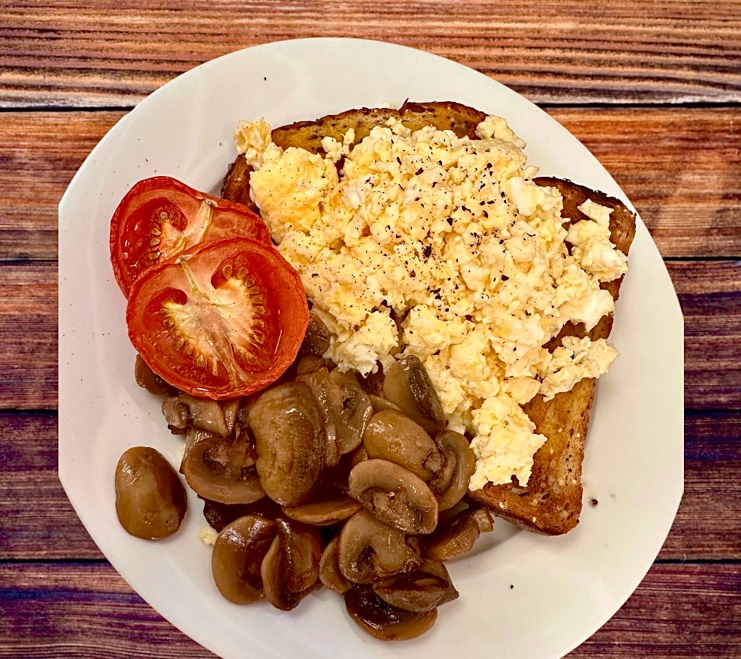 A plate is presented featuring toasted bread topped with scrambled eggs, accompanied by sautéed mushrooms and sliced tomatoes. The meal is arranged on a white dish against a backdrop of rustic wooden texture.