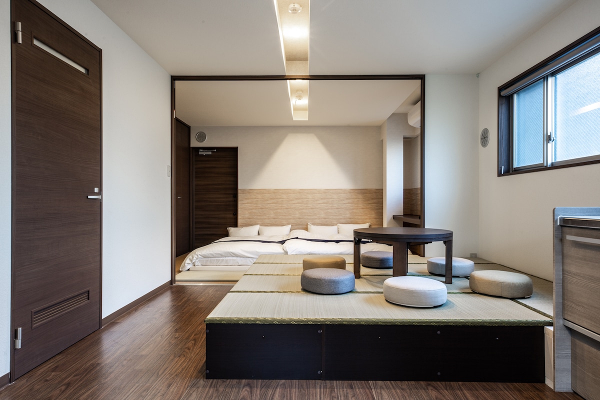A spacious room features a traditional tatami area with low seating and a round table. A sliding door reveals a sleeping area with neatly arranged futons. Natural light enters through large windows, enhancing the room's serene atmosphere.
