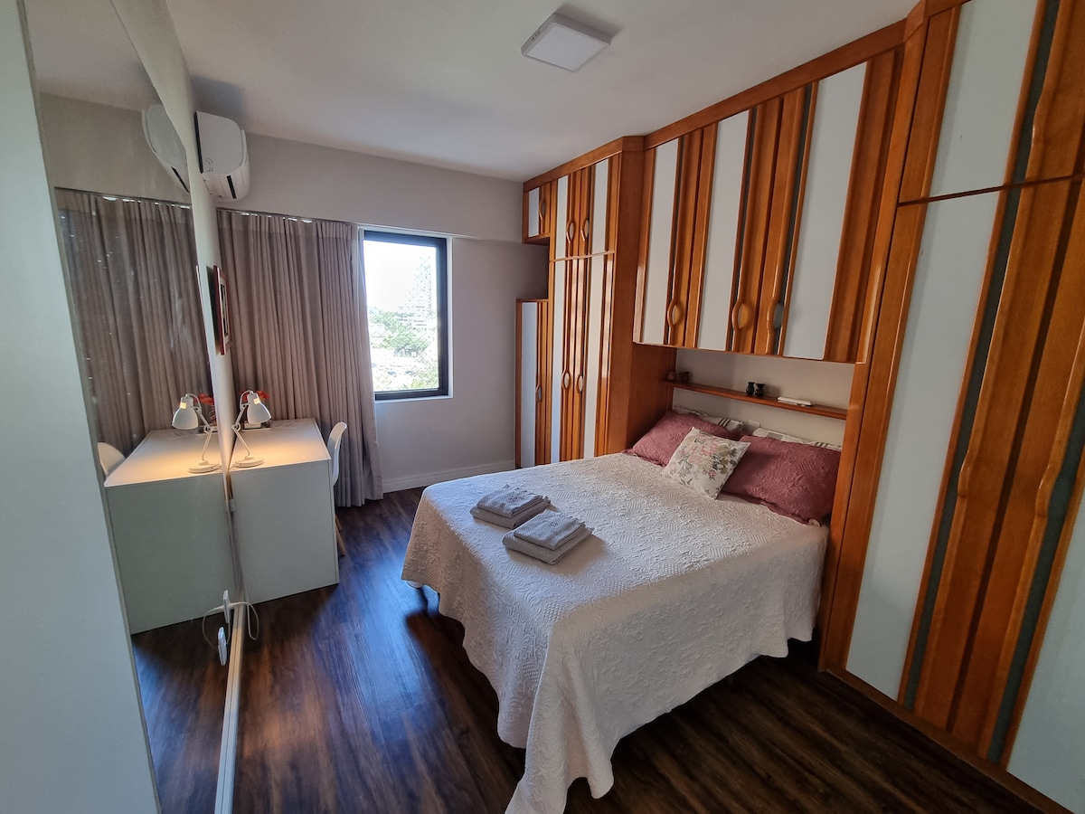 A well-appointed bedroom features a spacious bed with light-colored bedding and decorative pillows. A mirrored wardrobe reflects the room's modern design. Natural light enters through a window with sheer curtains, and a small desk with two lamps is positioned nearby.