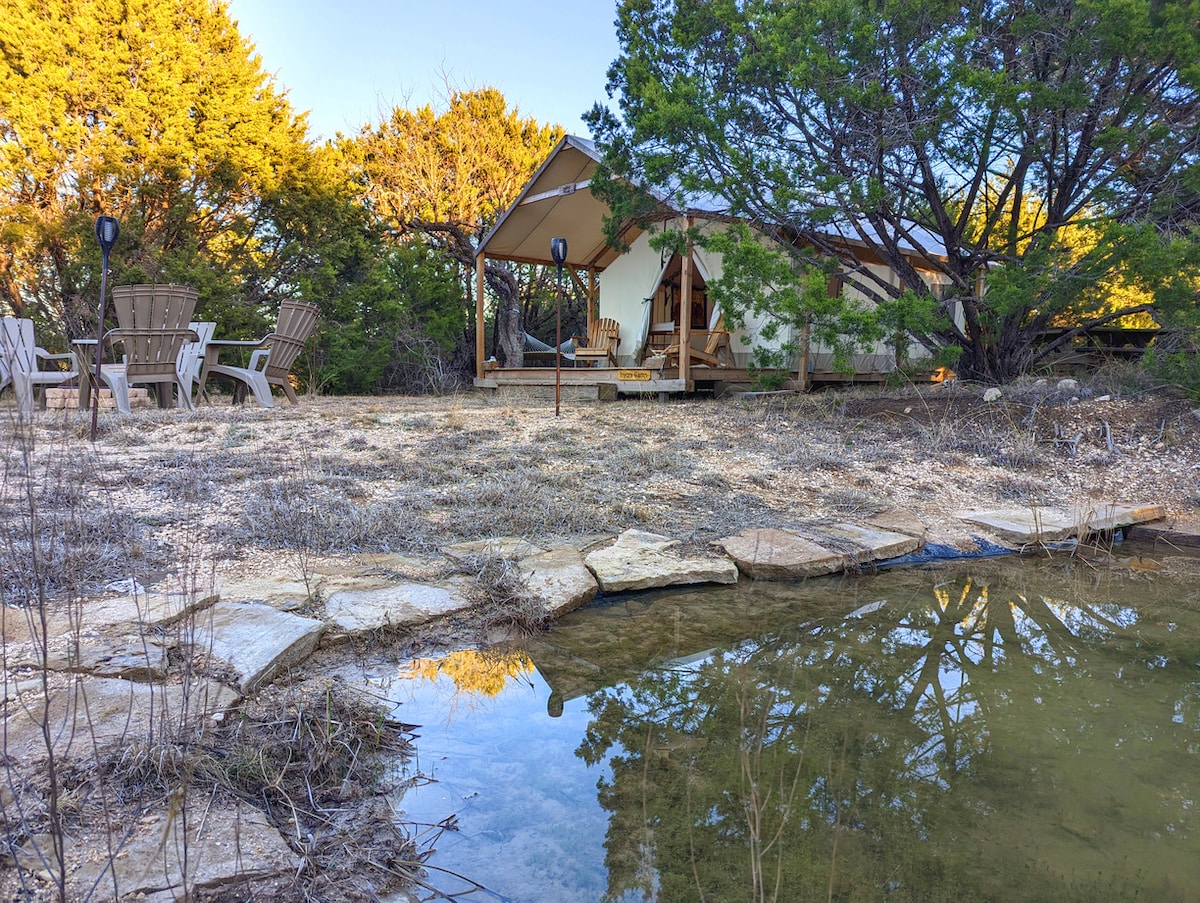 A safari tent is situated in a natural setting among trees, featuring a wooden platform and access steps. A small pond reflects surrounding vegetation, while several Adirondack chairs are arranged on gravel nearby, offering a space to relax and enjoy the outdoors.