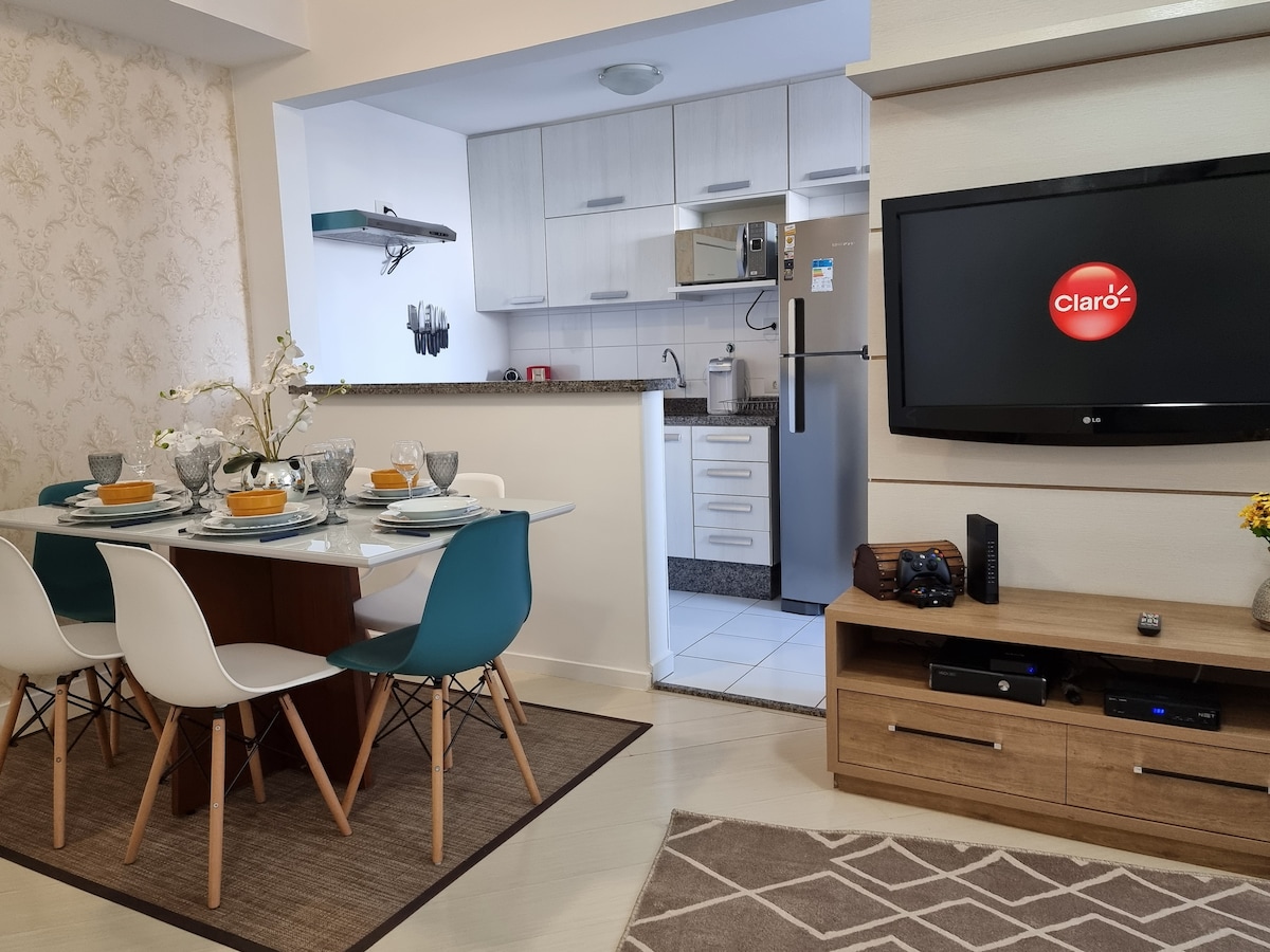 A modern dining area features a wooden table set for four, with blue and white chairs arranged around it. The adjoining kitchen is visible, displaying sleek cabinets and stainless steel appliances. A TV is mounted on the wall, with a textured rug adding warmth to the space.