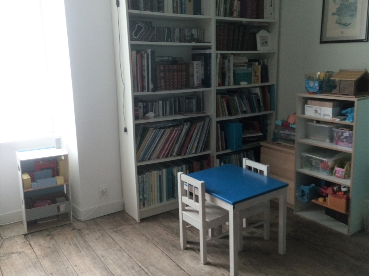 A children's playroom is furnished with a small table and two chairs, featuring a blue tabletop. Shelves are loaded with books and toys, alongside a light-colored storage unit. Natural light streams in from a nearby window, illuminating the room's wooden floor.