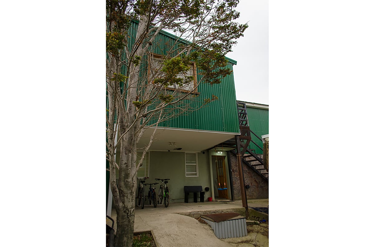 A green two-story building is visible with an exterior staircase leading to the upper level. In the foreground, two bicycles are parked alongside the entrance, which features a covered seating area. A tree is present, adding a natural element to the setting.