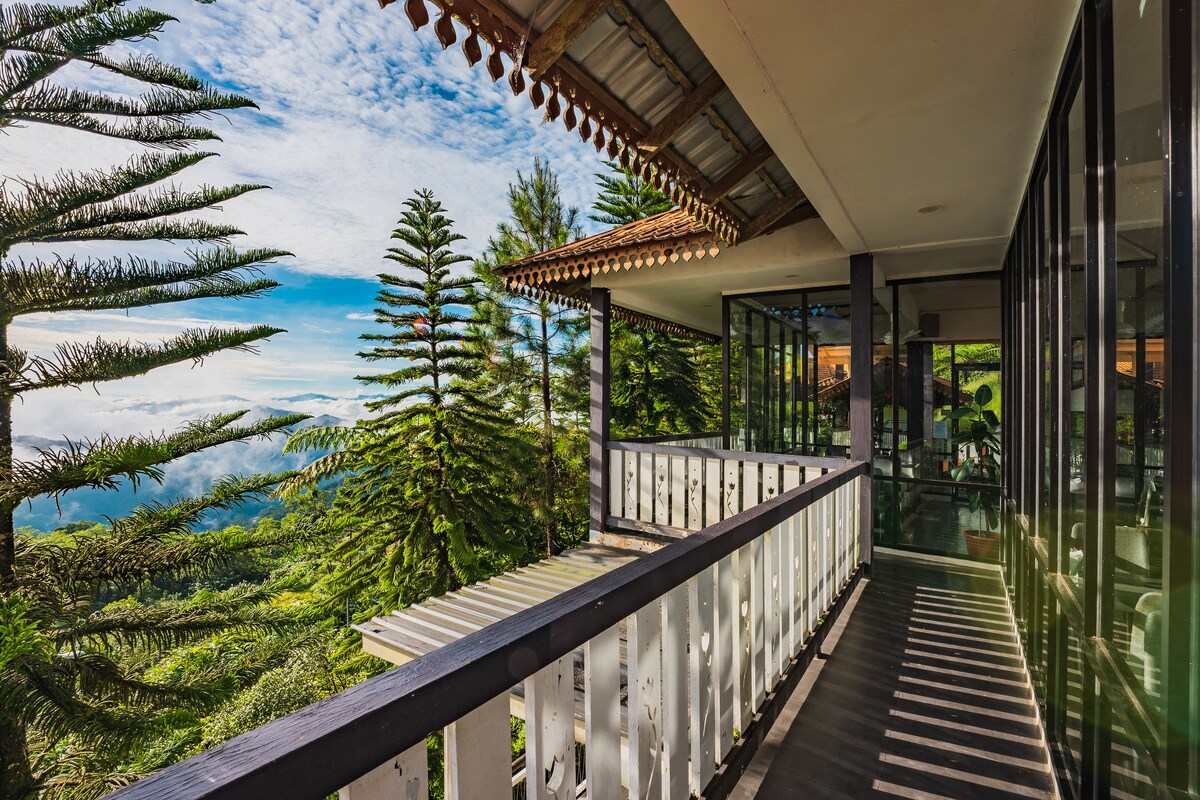 A spacious balcony offers views of surrounding lush greenery and distant mountains. Large glass doors provide access to the interior, while a wooden railing complements the natural setting. Sunlight casts shadows on the floor, highlighting the tranquil outdoor space.
