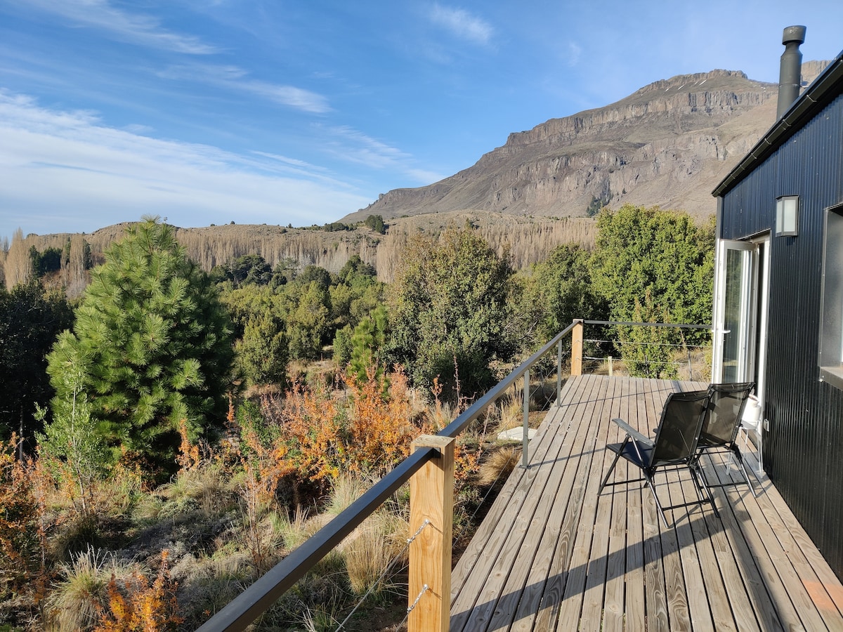 A spacious wooden deck is showcased, leading from the house and bordered by lush greenery. Views of a mountainous landscape are visible in the background, with a mix of vibrant foliage in warm tones. Two black chairs are positioned for relaxation, inviting guests to enjoy the outdoors.