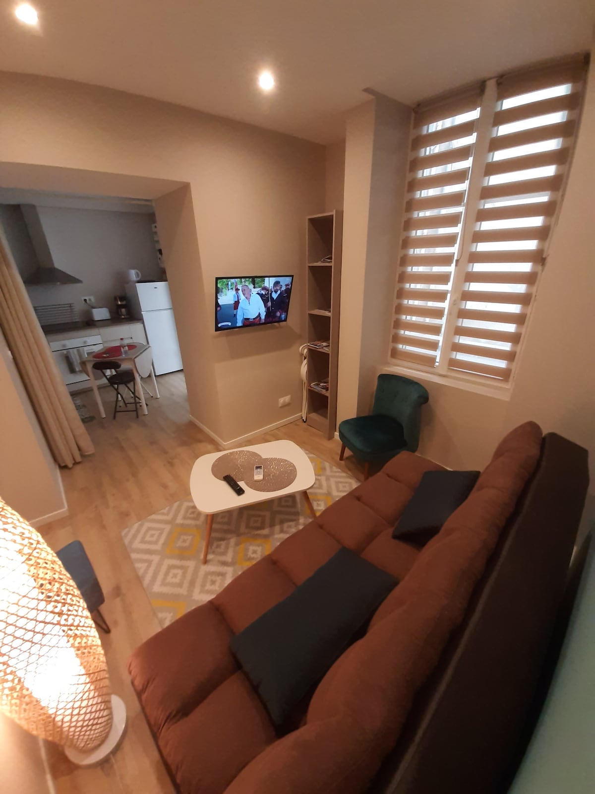 A cozy living area is presented with a brown sofa and blue accent pillows. A small coffee table is positioned on a patterned rug. A television is mounted on the wall, while a kitchen space is visible in the background. Light filters through the window covered by wooden shutters.