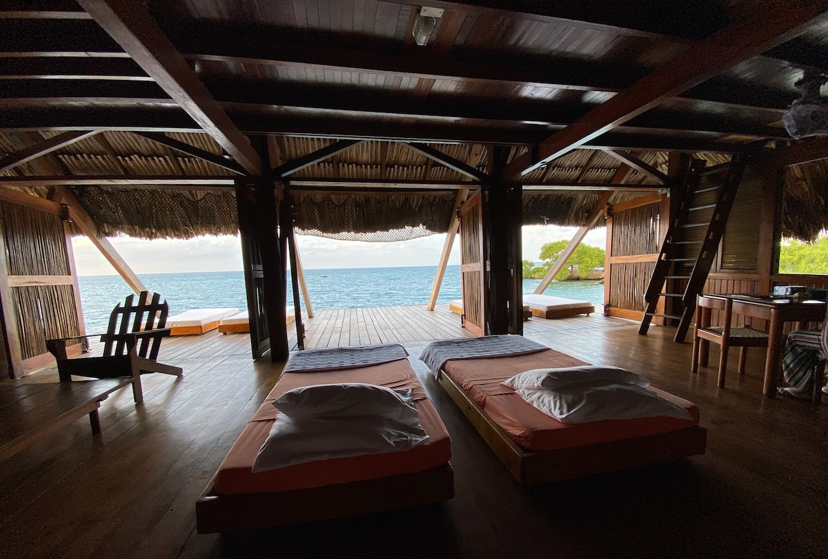 An open living space features two spacious beds with simple bedding, framed by large openings that provide a direct view of the sea. Natural light floods the room, accentuating the wooden beams and palm leaf roof. A cozy chair sits nearby, inviting relaxation.